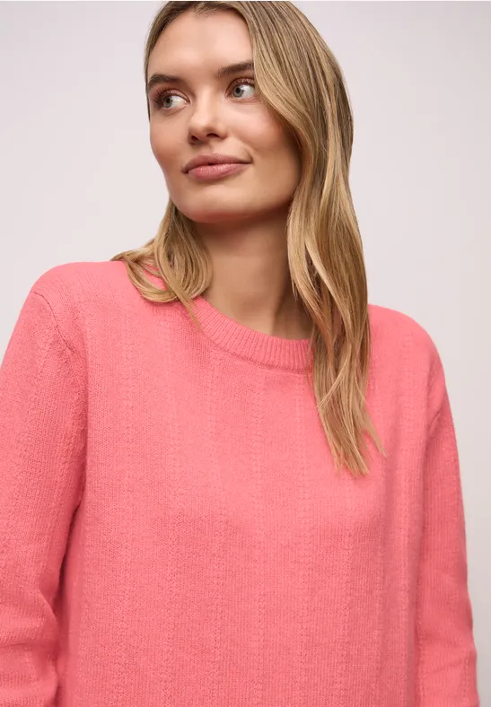 Frau mit langem blonden Haar in lockerem pinken Pullover vor hellem Hintergrund.