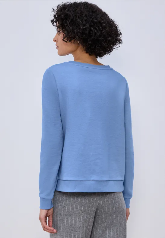 Frau von hinten in hellblauem Pullover und grauer Hose, braune Locken, heller Hintergrund.