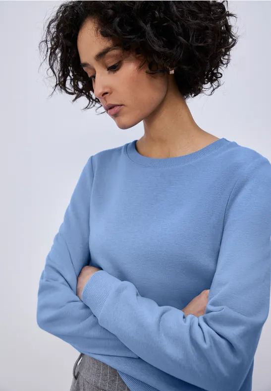Person in blauem Pullover mit verschränkten Armen vor neutralem Hintergrund.