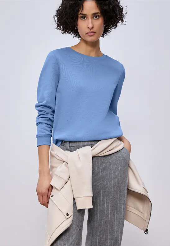 Person trägt hellblauen Pullover und graue Nadelstreifenhose, beige Jacke lässig um die Hüfte gebunden.
