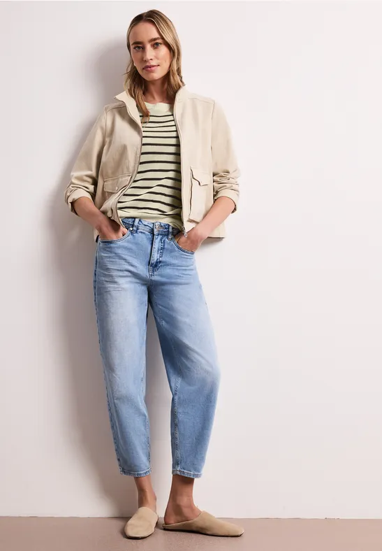 Eine Frau in lässigem Outfit: gestreiftes T-Shirt, beiger Blouson, weite Jeans und Slipper vor weißer Wand.