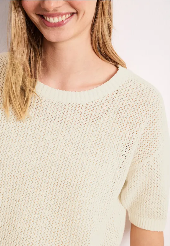Nahaufnahme einer lächelnden Frau in einem cremefarbenen, locker gestrickten Kurzarmpullover.