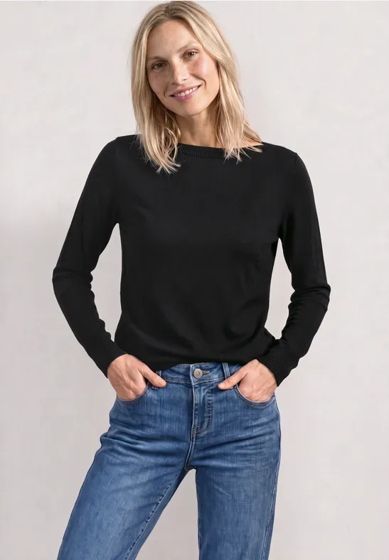 Frau in schwarzem Pullover und blauen Jeans, steht vor hellem Hintergrund mit Händen in den Taschen.
