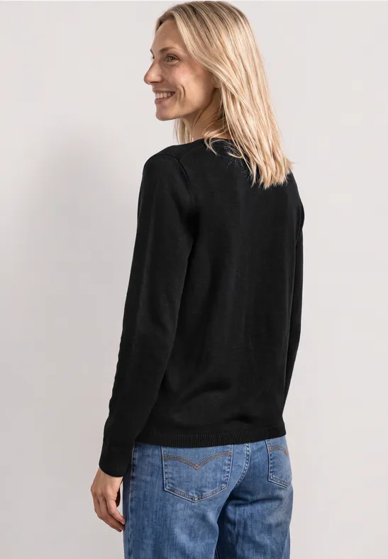 Frau mit blondem Haar trägt schwarzen Pullover und Jeans, Seitenansicht vor hellem Hintergrund.