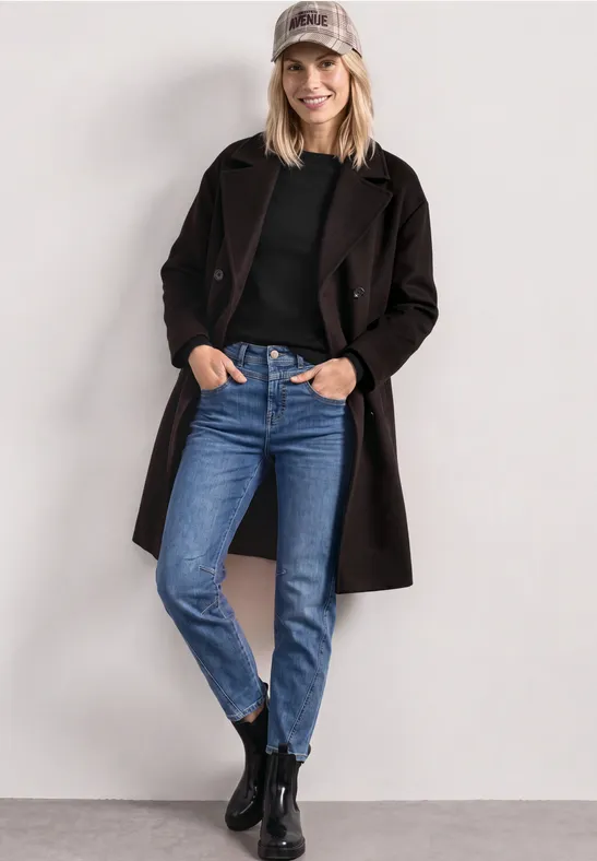 Junge Frau in Jeans, schwarzem Mantel, Baseballcap und Stiefeln, die lächelnd an eine Wand lehnt.