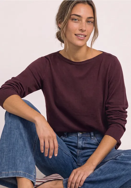 Une femme souriante en pull bordeaux et jean assise devant un fond blanc.