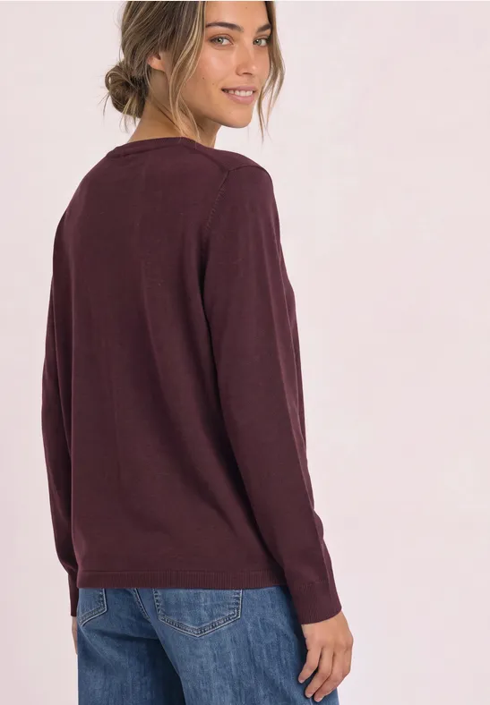 Une femme avec un chignon porte un pull bordeaux et un jean bleu, regard par-dessus l'épaule.