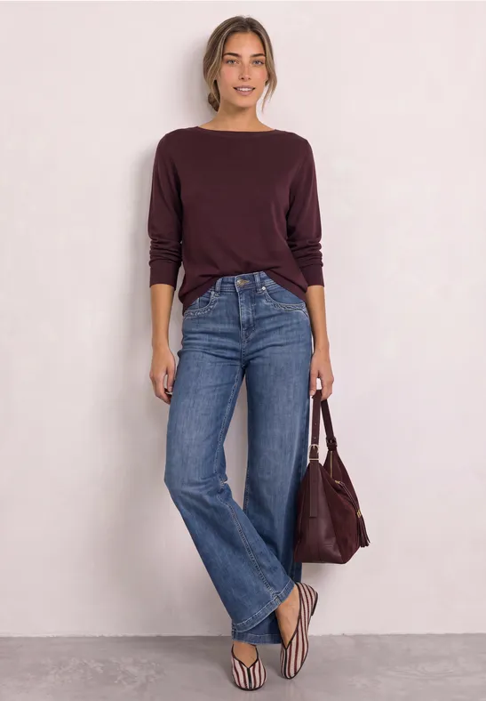 Femme en pull bordeaux, jean bleu large, tongs et sac assorti, posant devant un mur blanc.