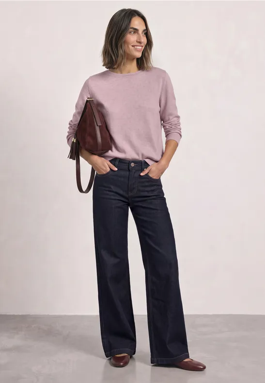 Eine Frau in rosafarbenem Pullover, weiten Jeans und braunen Schuhen mit einer roten Handtasche steht lässig.