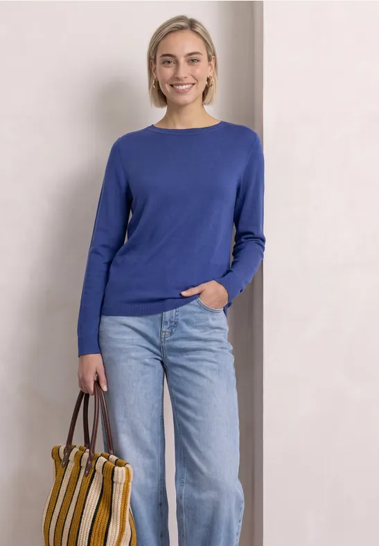 Eine Frau steht vor hellem Hintergrund und trägt einen blauen Pullover, Jeans und eine gestreifte Tasche.