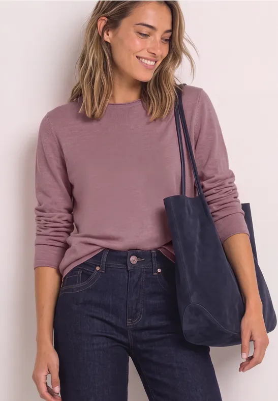 Vrouw lacht, draagt een roze trui, donkerblauwe jeans en heeft een blauwe tas over de schouder.