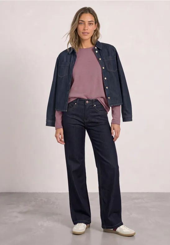 Vrouw met roze trui, wijd donkerblauw jeans, spijkerjasje over schouders en witte sneakers.