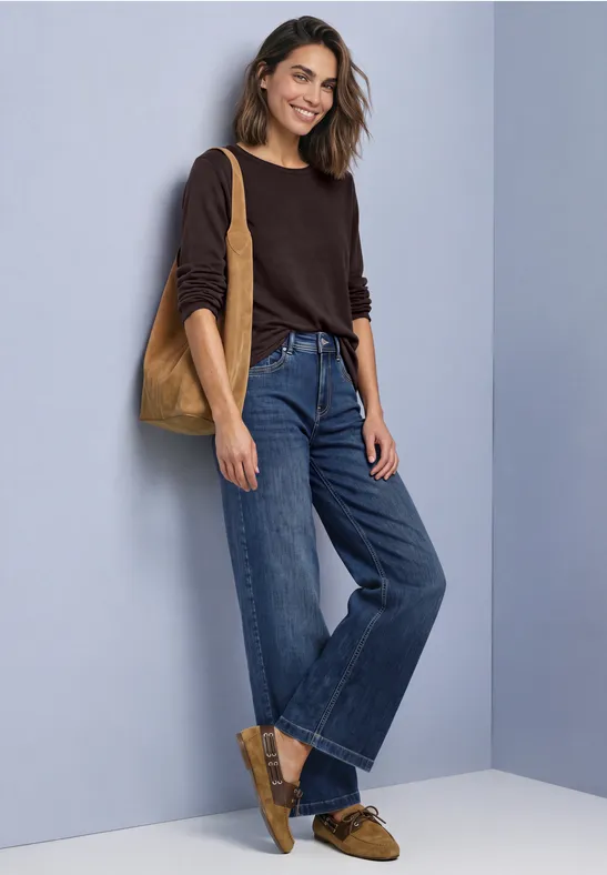 Lachende vrouw in bruine trui, wijde jeans en bijpassende loafers met tas voor een grijze achtergrond.