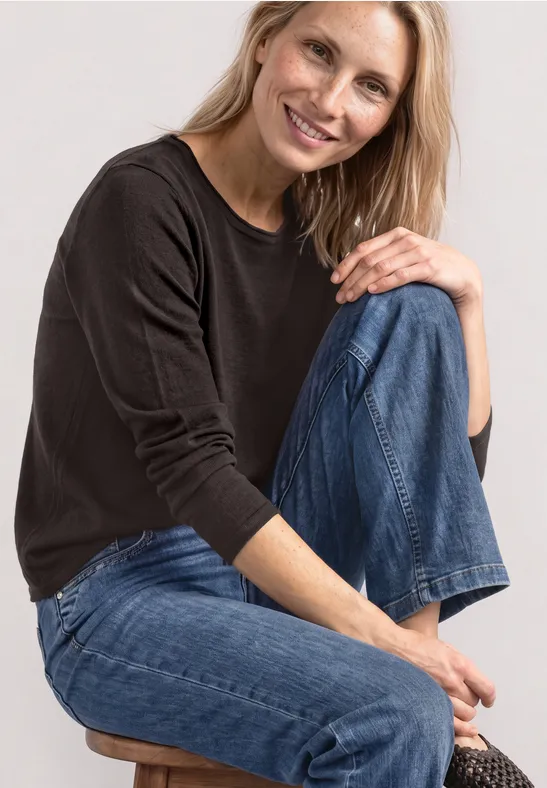 Een glimlachende vrouw in zwarte trui en blauwe jeans zit op een kruk.
