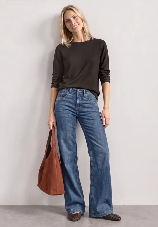 Vrouw in zwarte trui, wijd blauwe jeans en bruine tas lacht voor een neutrale achtergrond.