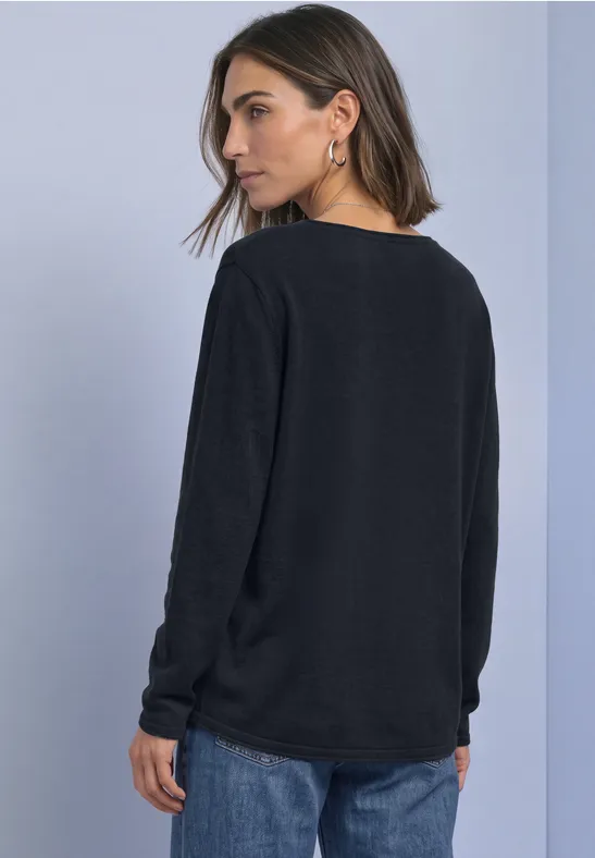 Vrouw in zwarte trui en blauwe jeans, zijwaarts gewend tegen lichte achtergrond.