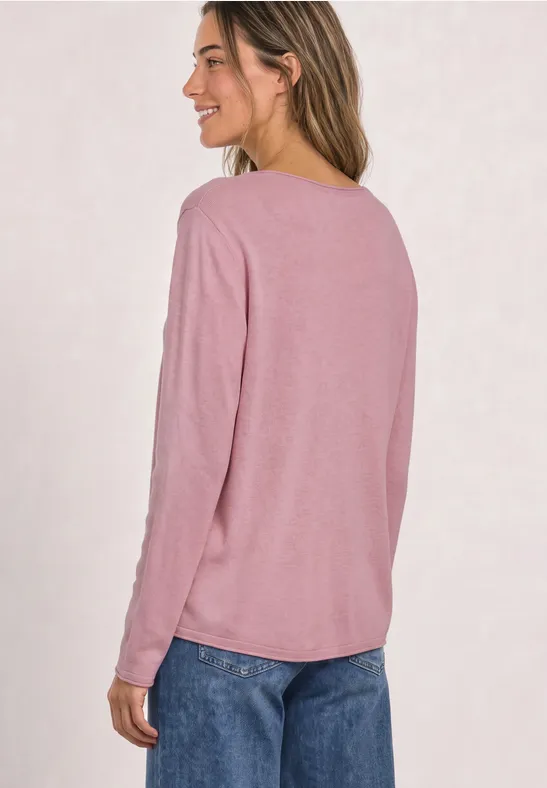 Een vrouw in roze langarmtrui en blauwe spijkerbroek, zijwaarts pose, glimlacht.