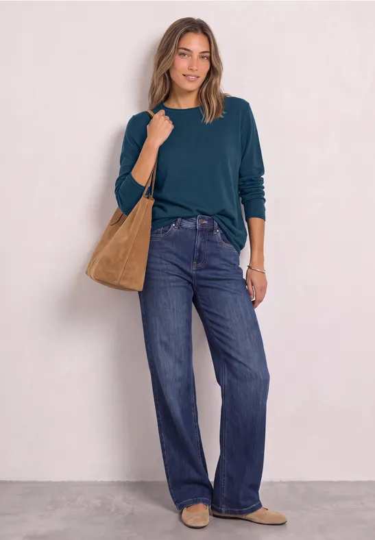 Een vrouw draagt een donkergroene longsleeve, wijde jeans, een bruine schoudertas en bijpassende schoenen.