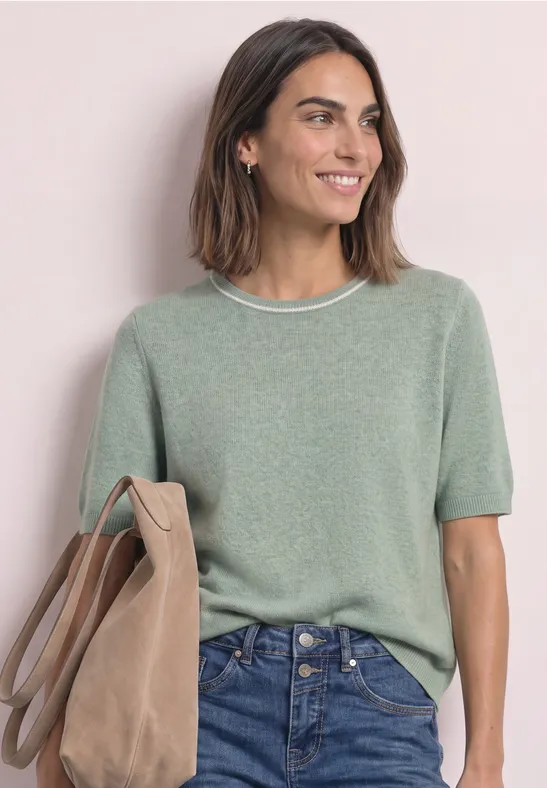 Femme souriante en pull vert à manches courtes, jean et sac beige devant un fond clair.