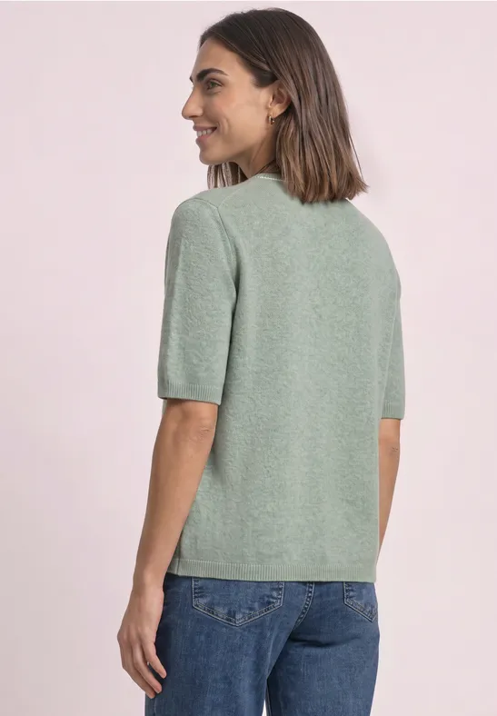 Femme de dos en pull vert clair à manches courtes et jean bleu, regard vers la droite sur fond clair.