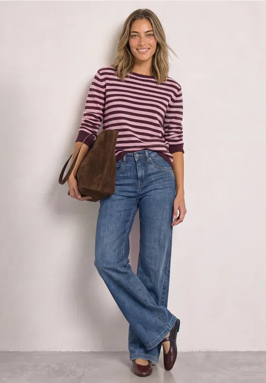 Frau im gestreiften Pullover, weiten Jeans und braunen Loafers trägt eine braune Tasche und lächelt.