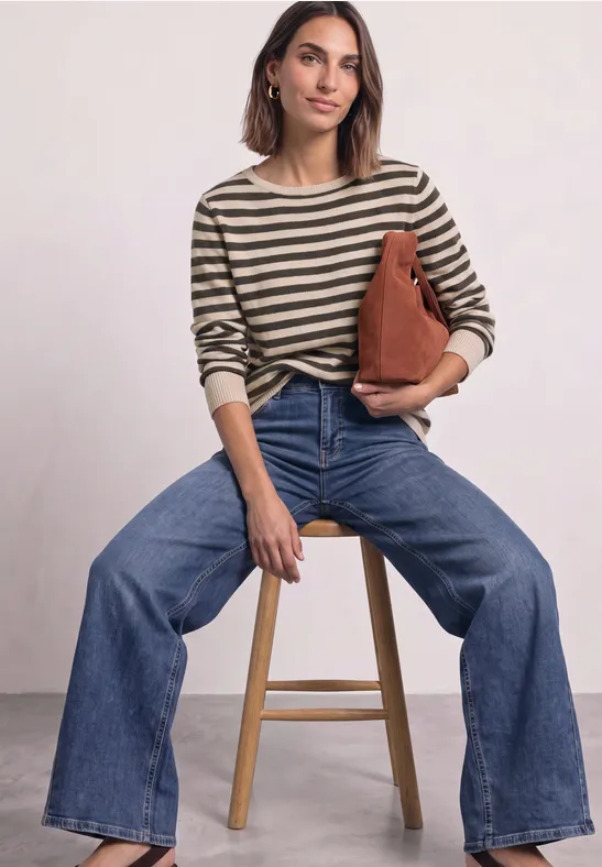 Femme assise sur un tabouret, porte un pull à rayures, un jean flare et tient un sac marron.