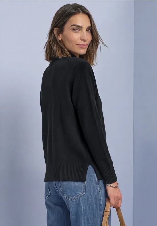 Frau in schwarzem Pullover und Jeans schaut über die Schulter vor hellviolettem Hintergrund.