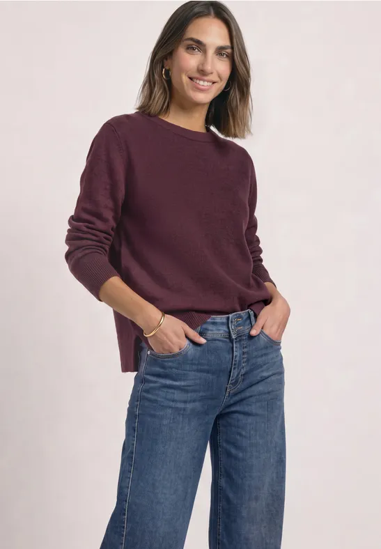 Eine lächelnde Frau trägt einen bordeauxfarbenen Pullover und blaue Jeans.