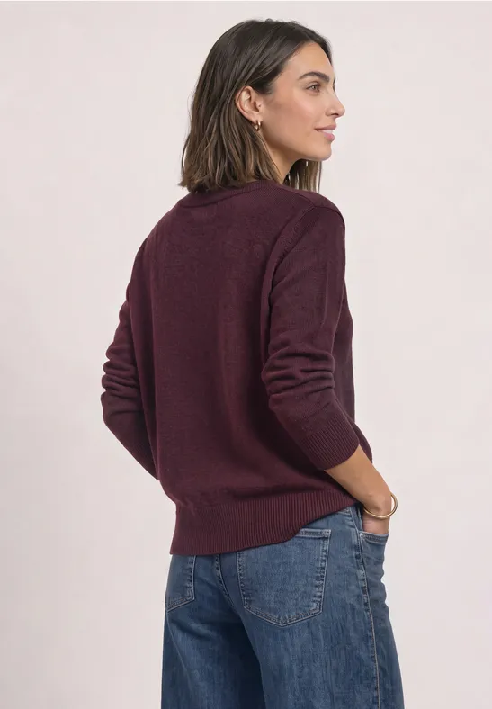 Frau von hinten in weinrotem Pullover und Jeans, linker Arm locker in Tasche.