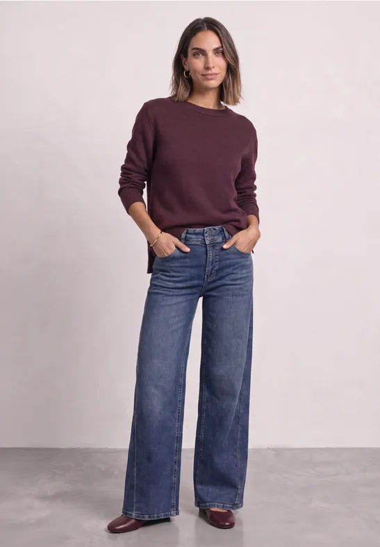 Frau in weinrotem Pullover und weiten Jeans, steht lässig mit Händen in den Taschen.