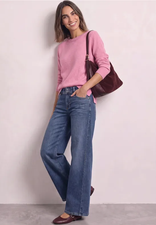 Jeune femme souriante vêtue d'un jean ample et d'un pull rose, portant un sac à épaule marron.