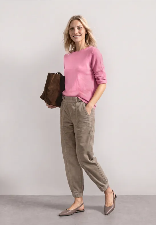Frau in rosa Pullover, khakifarbener Cargo-Hose und brauner Tasche steht vor hellem Hintergrund.