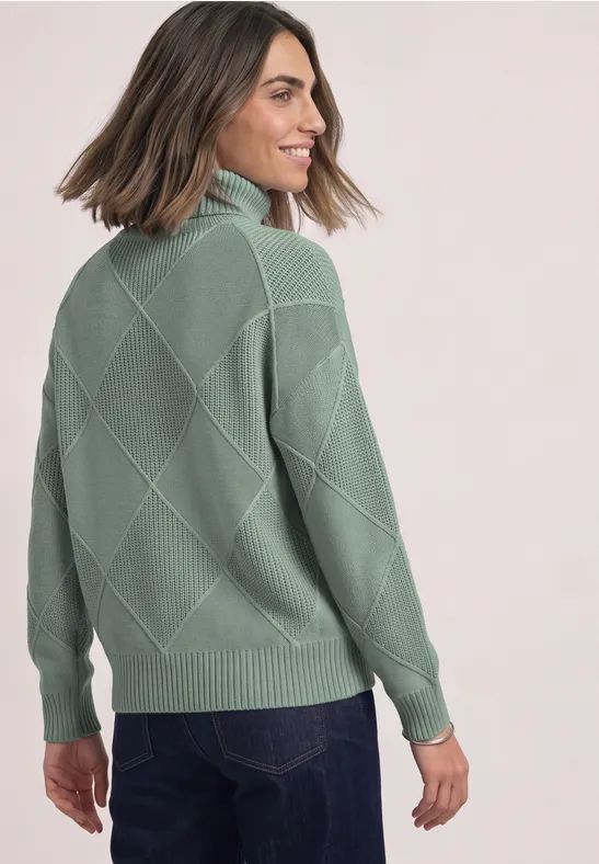 Frau sieht über die Schulter, trägt grünen Strickpullover mit Rautenmuster und Jeans.