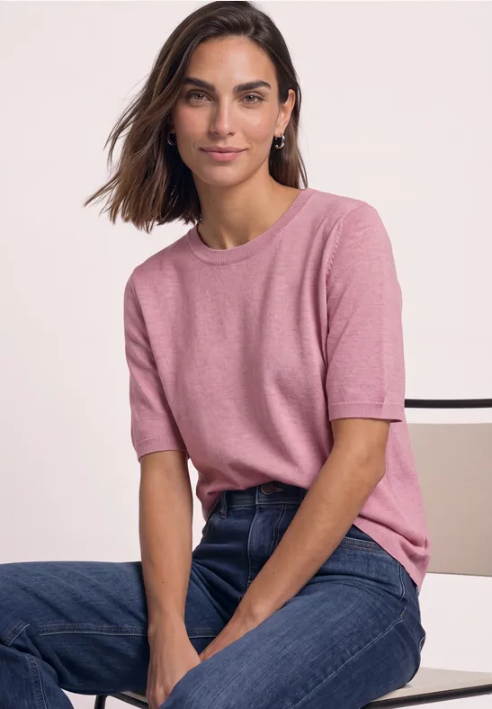 Frau mit rosa T-Shirt und Jeans sitzt, lächelt in die Kamera.