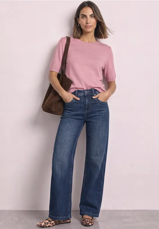 Frau in rosa T-Shirt und weiten Jeans, trägt eine braune Tasche, steht vor hellem Hintergrund.