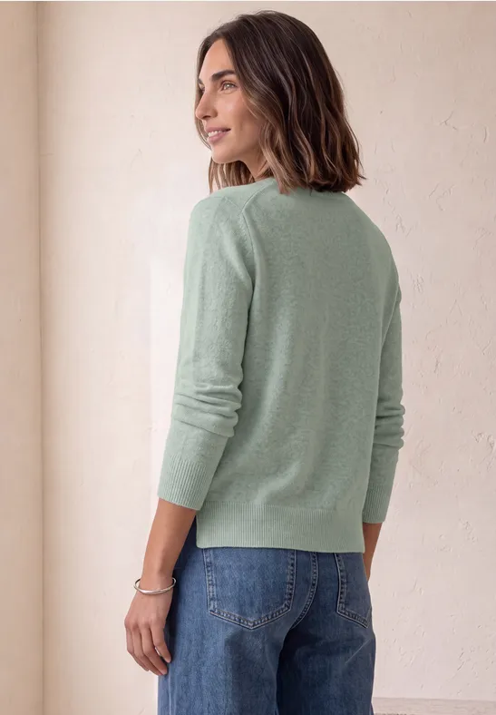 Une femme de profil porte un pull vert clair et un jean bleu, regardant par-dessus son épaule sur un fond neutre.