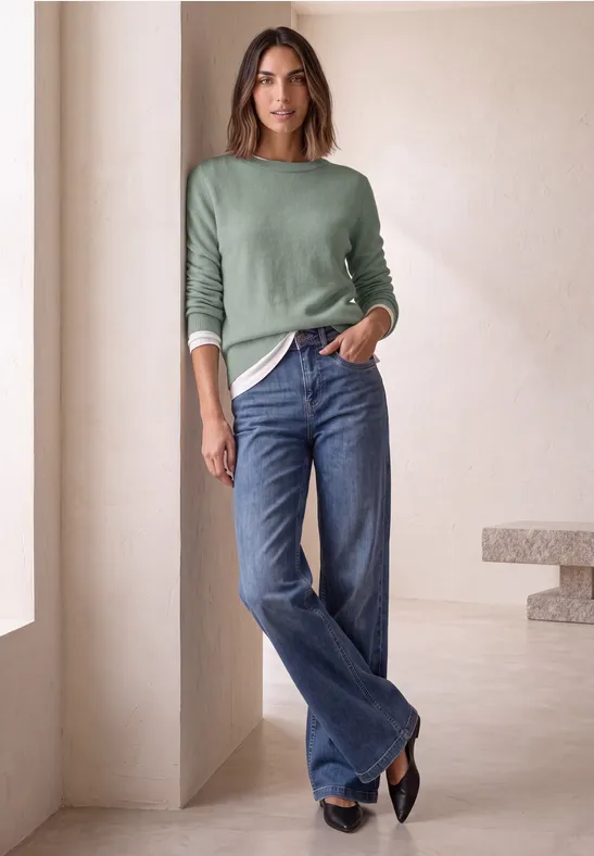 Femme en pull vert et jean large, posant détendue dans un intérieur lumineux et minimaliste.