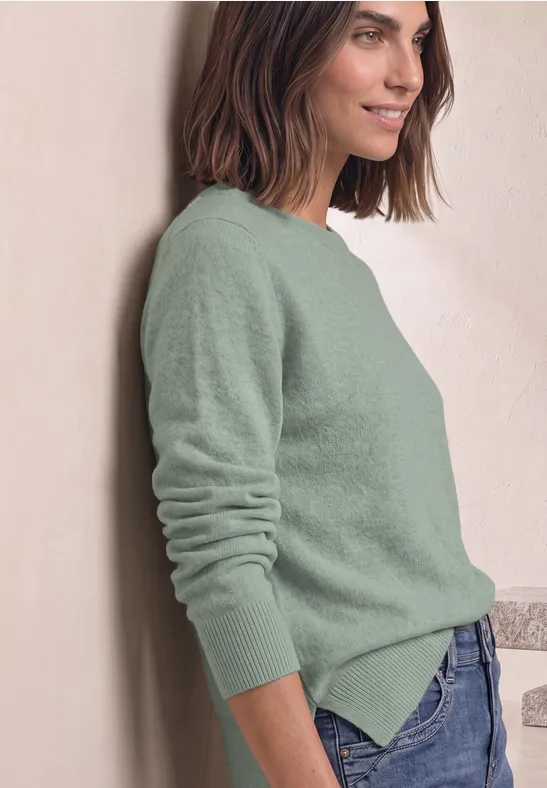 Femme appuyée contre un mur, pull vert et jean bleu, sourire détendu.