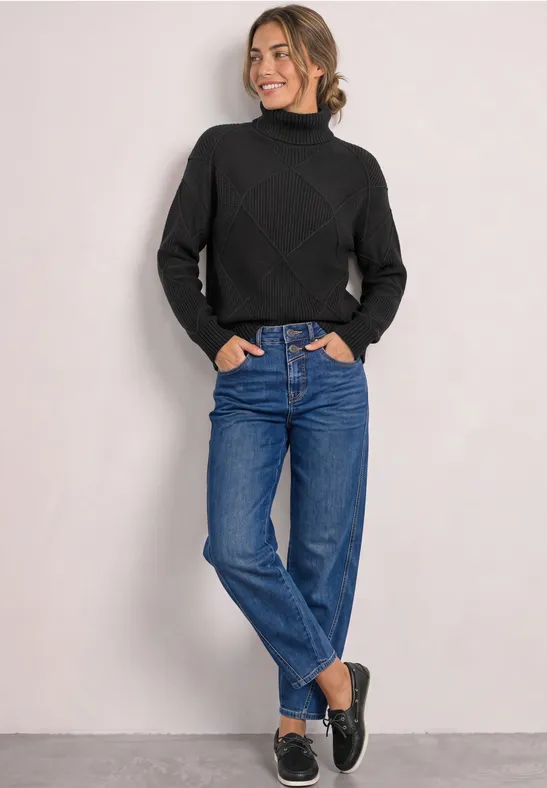 Jonge vrouw in zwarte coltrui en blauwe jeans voor een neutrale achtergrond.