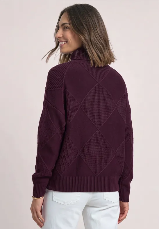 Vrouw van achteren in paarse gebreide trui met opstaande kraag en ruitpatroon, witte broek, glimlacht opzij.