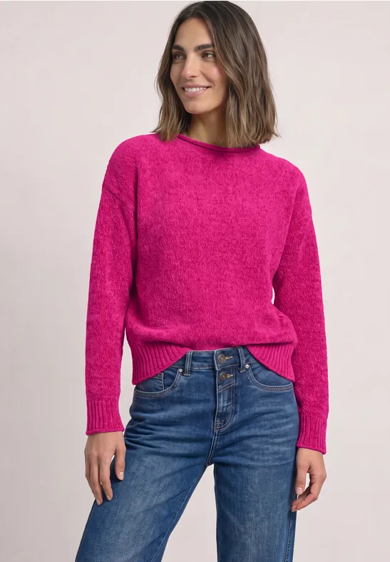Vrouw lachtend in een roze trui en blauwe jeans voor een lichte achtergrond.