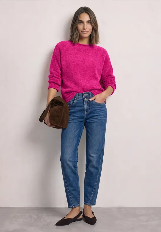 Vrouw in een roze trui, blauwe jeans en zwarte ballerina’s, met een bruine clutch, staand voor een lichte achtergrond.