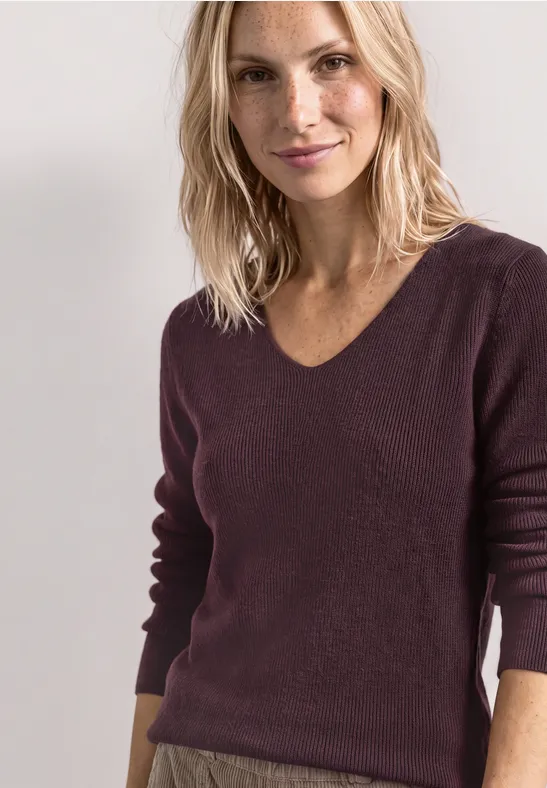 Blonde Frau im bordeauxfarbenen Pullover vor hellem Hintergrund, lächelnd und lässig posierend
