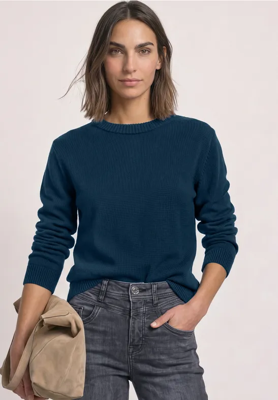 Frau in marineblauem Pullover und grauer Jeans, hält eine beige Jacke, vor hellem Hintergrund.