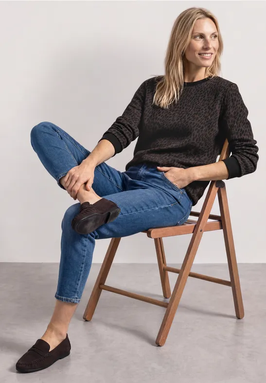 Une femme assise, jambes croisées sur une chaise en bois, pull, jean et mocassins, souriante.