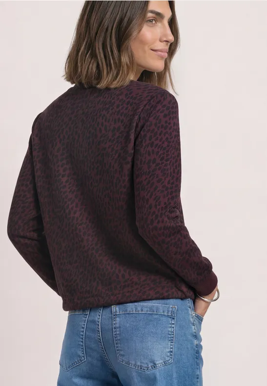 Femme vue de côté en pull violet à motif léopard et jean bleu, cheveux mi-longs, regard par-dessus l’épaule.