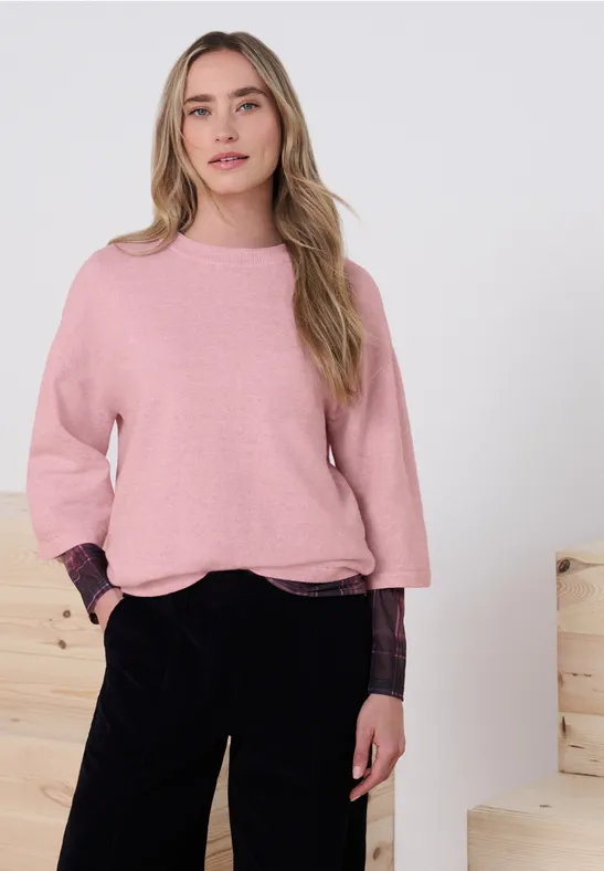 Jeune femme porte un pull rose et un pantalon noir, mains dans les poches, devant des blocs de bois clairs.