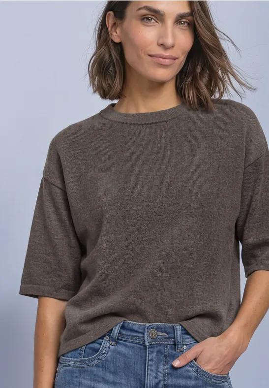 Femme en pull brun à manches courtes et jean bleu, main dans la poche, posant devant un fond clair.