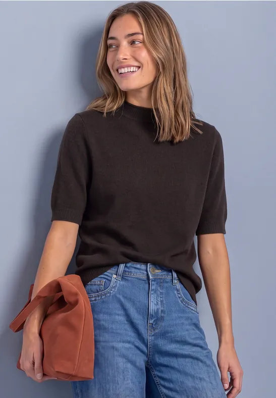 Lächelnde Frau im schwarzen Kurzarm-Pullover hält eine rostrote Tasche, steht vor grauem Hintergrund.