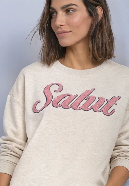 Femme portant un sweat clair avec l'inscription rose 'Salut', souriante devant un fond neutre.